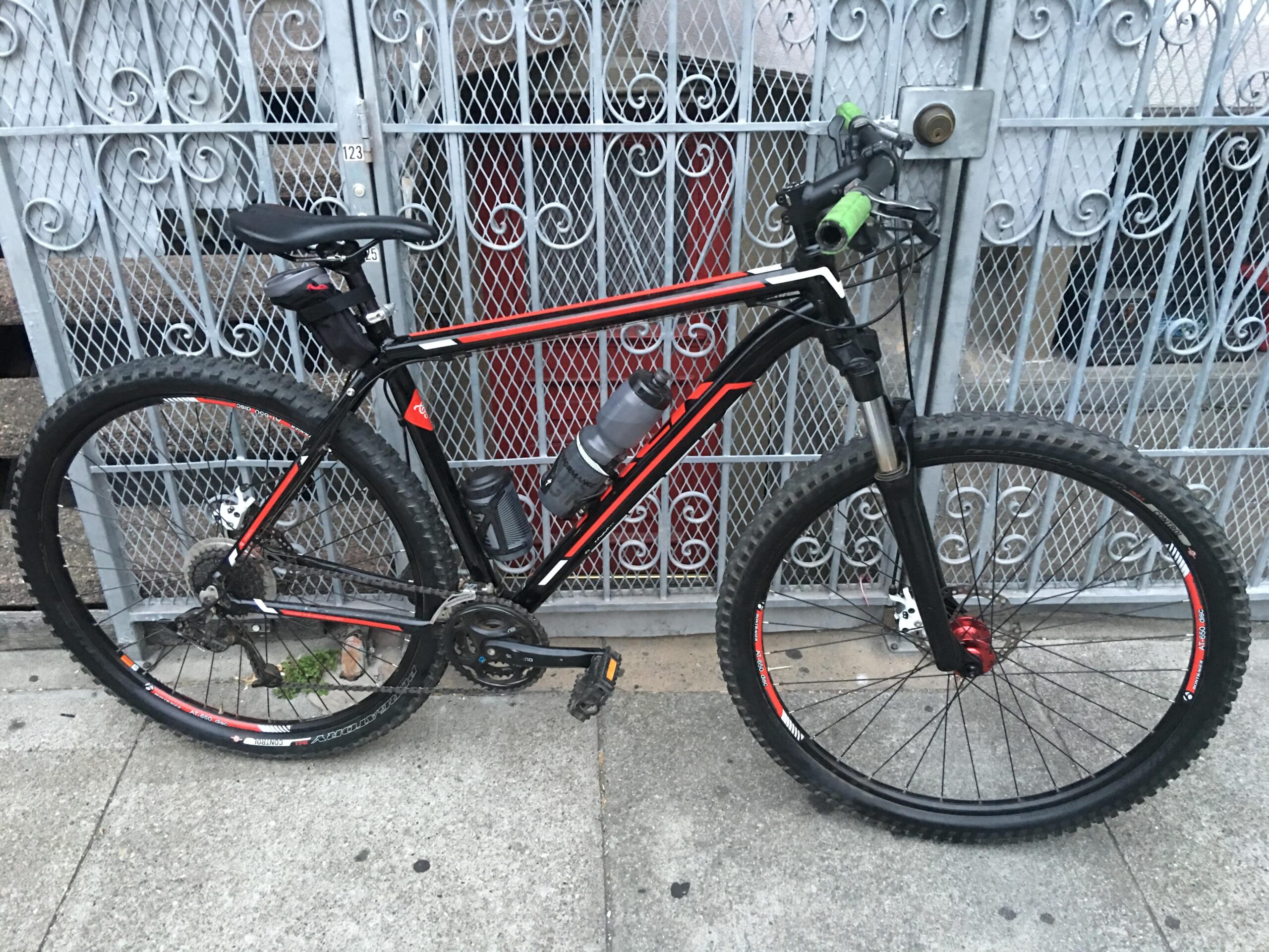 Trek Marlin 5 29er: A black and red mountain bike is parked against a decorative metal gate with a lock. The bike features thick tires, a water bottle mounted on the frame, and green handlebar grips. The ground is paved, and some greenery can be seen near the bike's rear wheel.
