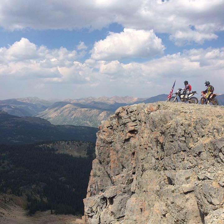 Two dirt bikers stand on the edge of a rocky cliff, overlooking a vast mountainous landscape with rolling hills and a cloudy sky. One biker is holding a U.S. flag, while the other is wearing a helmet. The scene captures a sense of adventure and exploration in a rugged outdoor setting. Flag mountain bike trail.