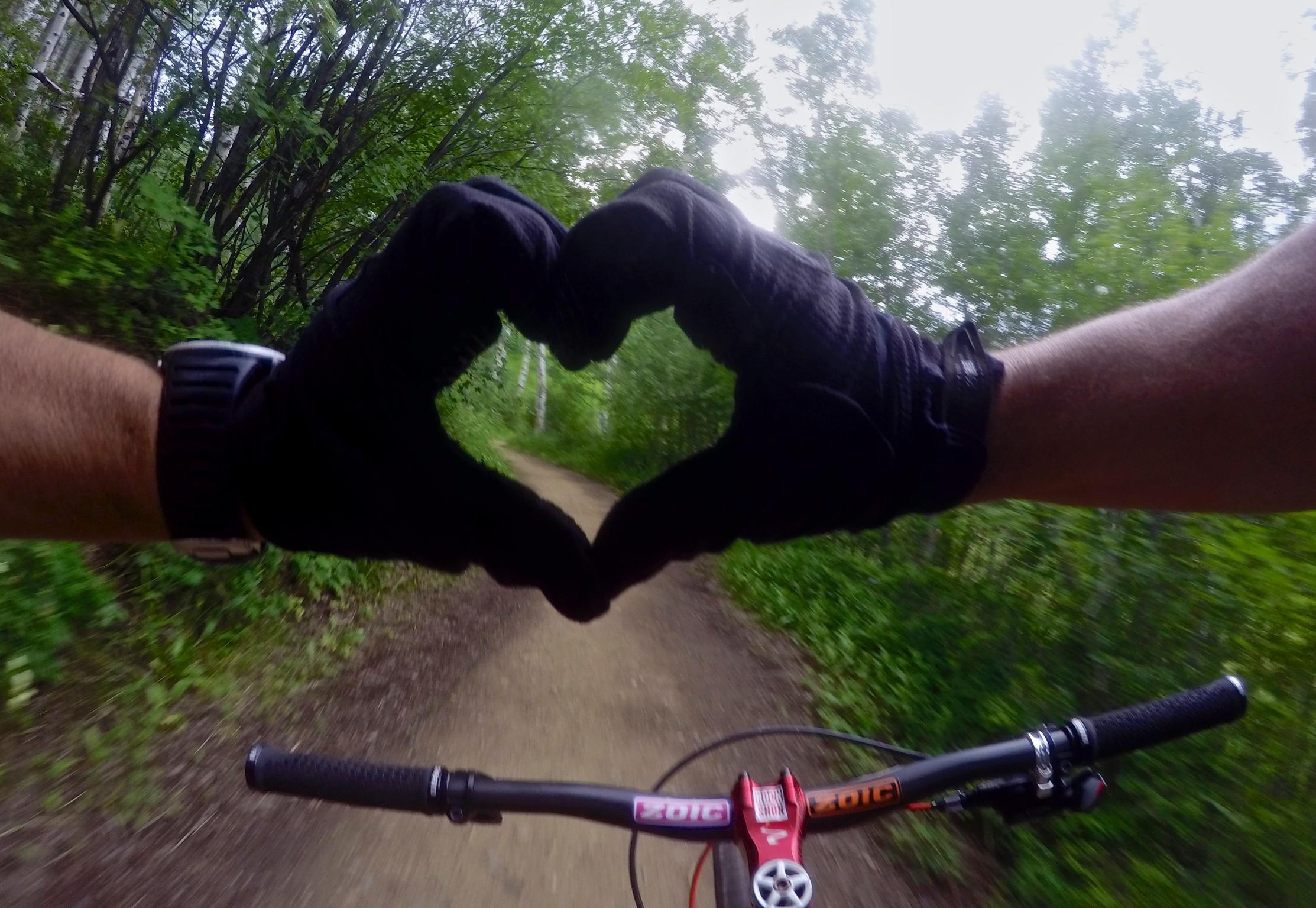 An image of a mountain biking scene where two gloved hands are forming a heart shape against a backdrop of a green, tree-lined dirt path, with the bike handlebars visible in the foreground. Beaver Creek Ski Resort mountain bike trail.
