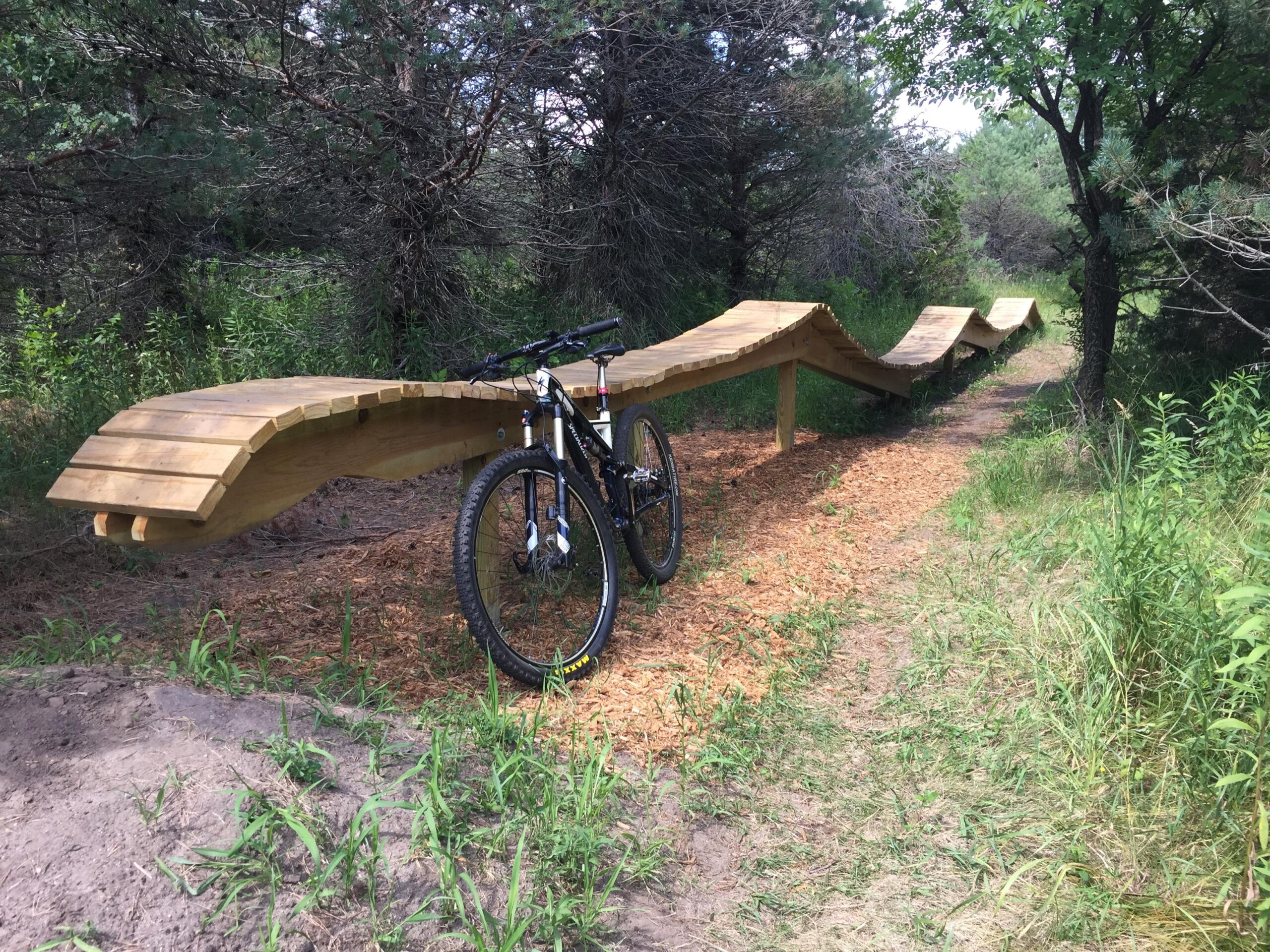 A mountain bike rests beside a wooden trail feature that resembles a series of undulating waves, surrounded by lush greenery and trees. The pathway is lined with crushed bark, leading into a natural setting. Bertram Chain of Lakes Trail mountain bike trail.