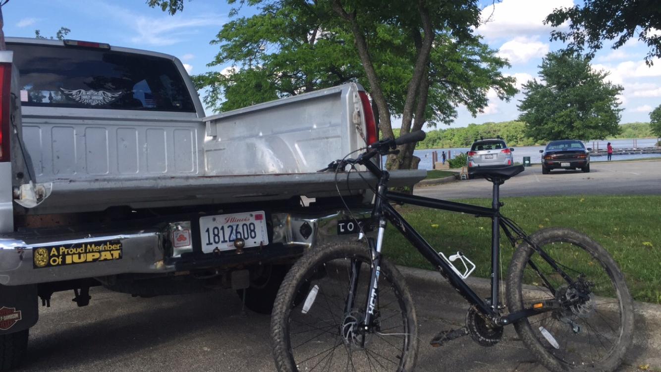 Nashbar AT-2: A black mountain bike is parked beside a gray pickup truck with a visible Illinois license plate, in a scenic area near a body of water. The background features trees and a few cars parked next to the water, with people enjoying the outdoors.