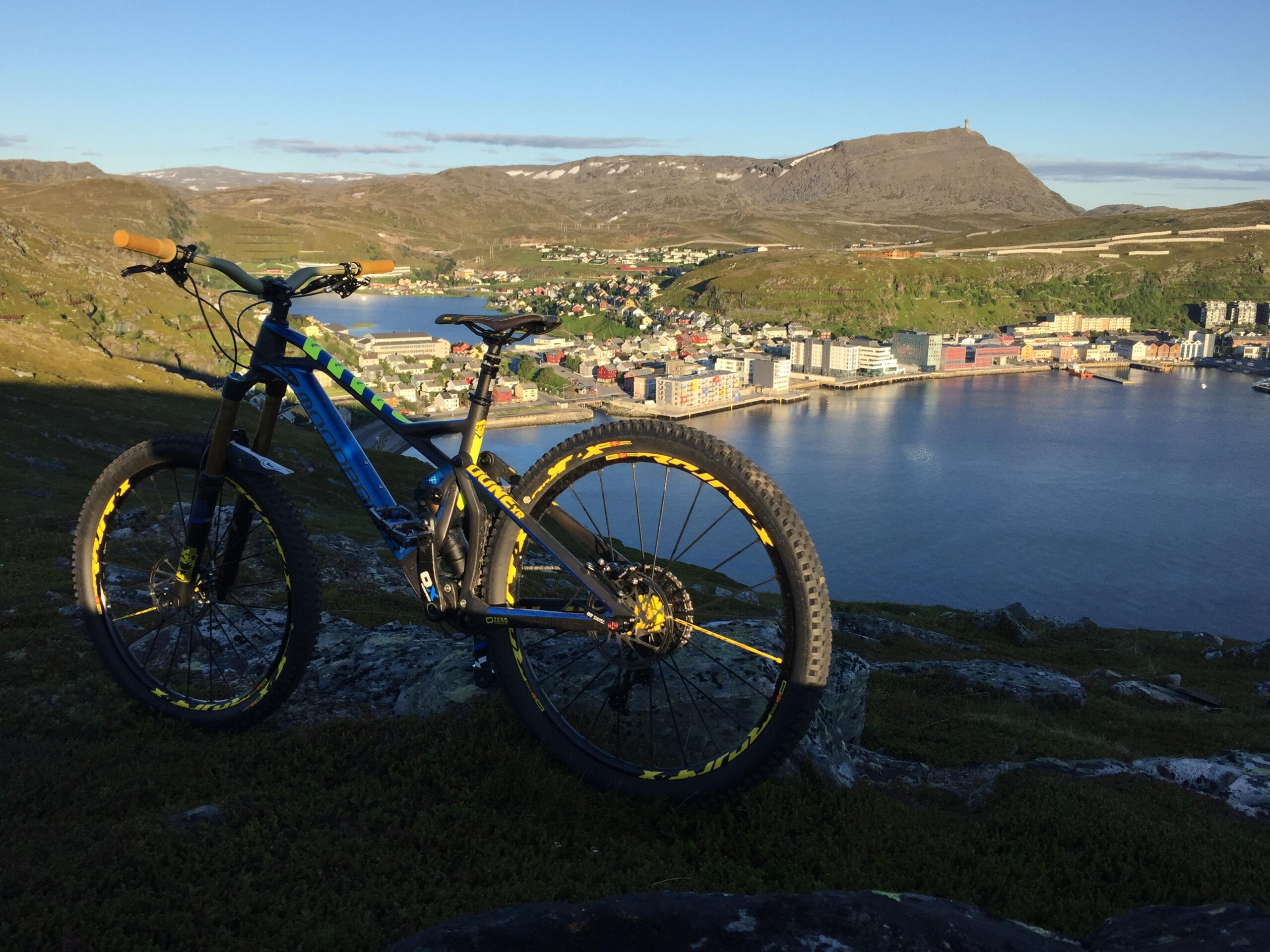 Mondraker Dune XR: A mountain bike positioned on rocky terrain overlooking a scenic coastal town and a calm body of water, with mountains in the background. The scene is bathed in natural sunlight, highlighting the vibrant colors of the bike and the surrounding landscape.