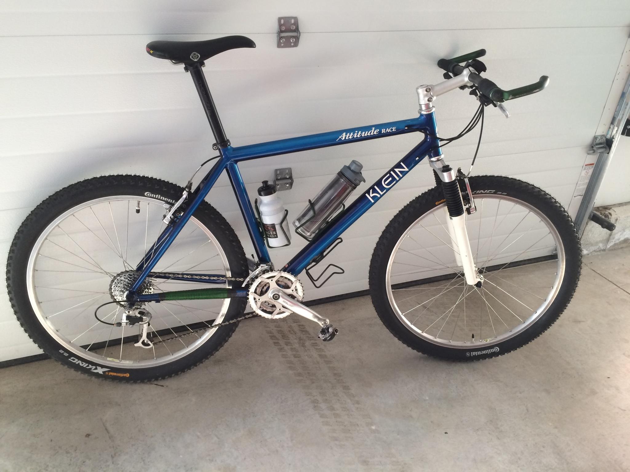 Klein Race: A blue mountain bike with a sleek design, featuring curved handlebars, a black seat, and a water bottle attached to the frame. The bike is positioned on a concrete floor near a garage door. The wheels are equipped with all-terrain tires, and the bike has a white front fork. The brand "Klein" is prominently displayed on the frame.