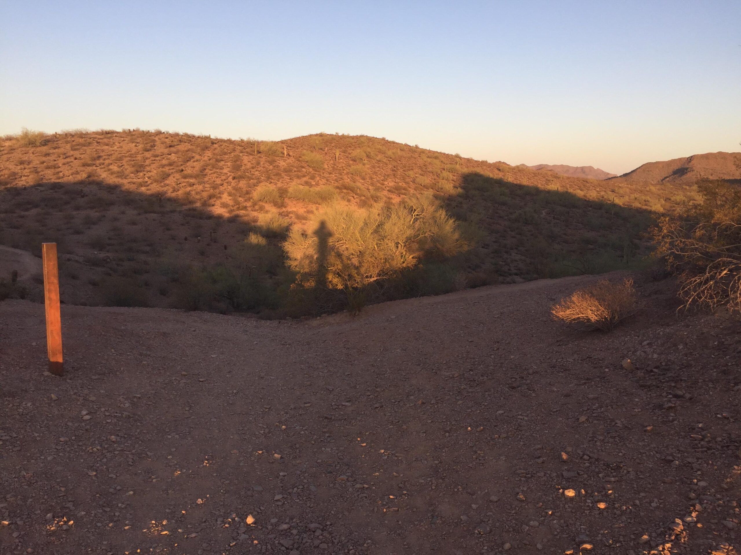 A pathway leading through a desert landscape at sunset, with gentle hills and sparse vegetation. A wooden post stands to the left, casting a long shadow on the uneven terrain. The warm hues of the setting sun illuminate the hills, creating a serene atmosphere. Trail #100 mountain bike trail.