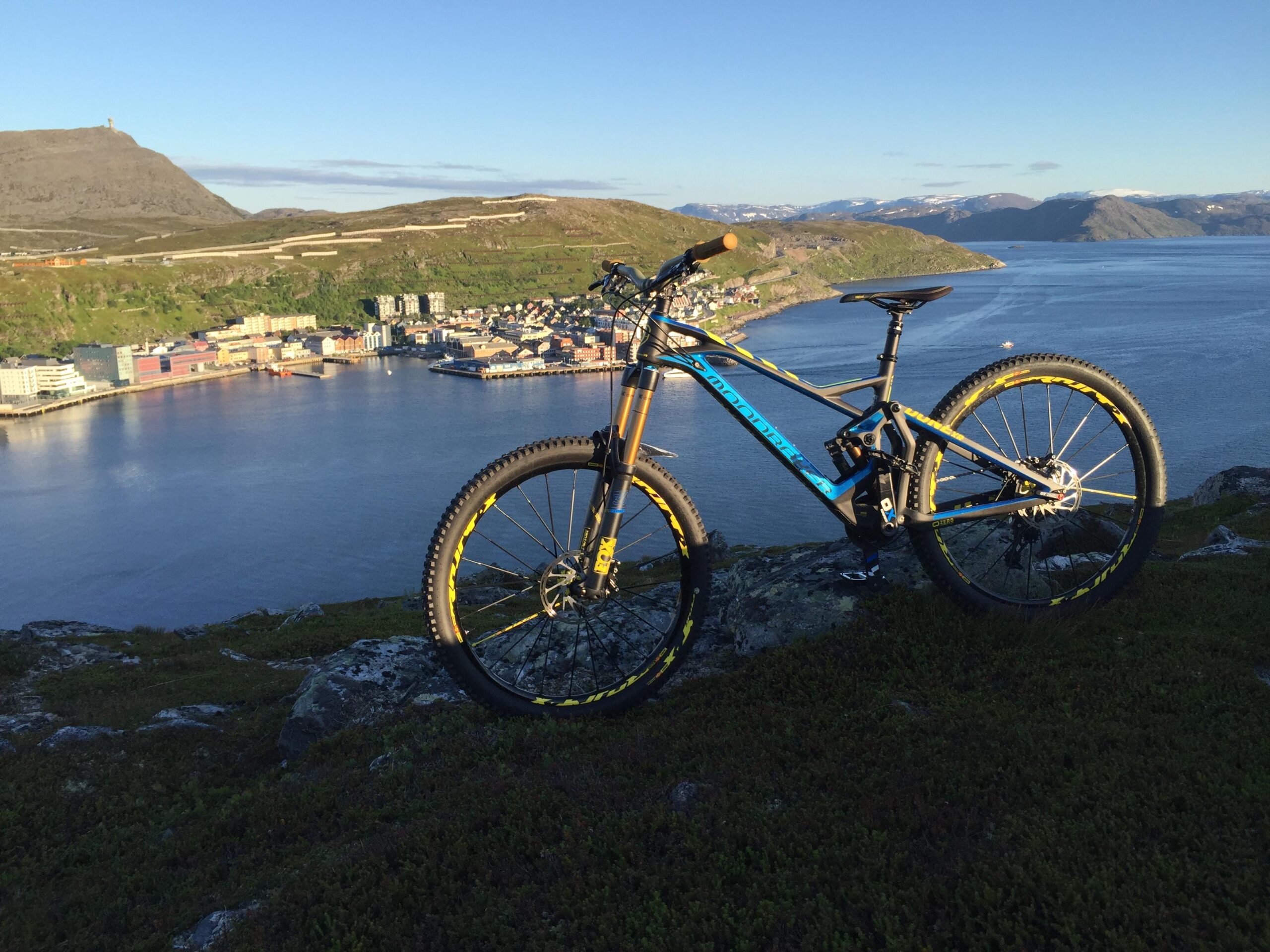 Mondraker Dune XR: A mountain bike positioned on a rocky outcrop overlooking a scenic coastal town and a calm body of water. The backdrop features lush green hills and a clear blue sky, with distant snow-capped mountains visible in the background.