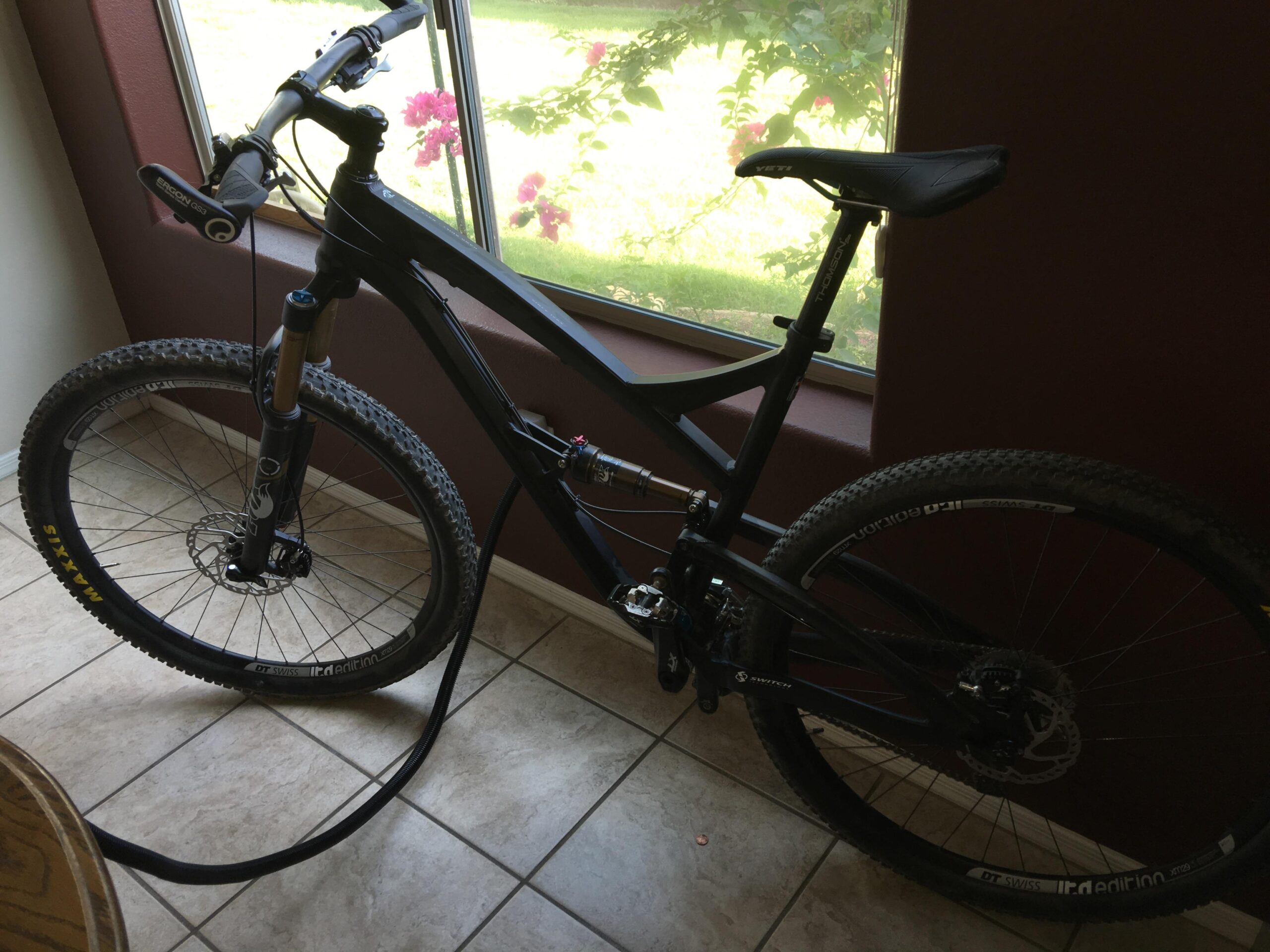 Yeti SB95: A black mountain bike resting on tiled flooring near a window, with a view of greenery and flowers outside. The bike features thick, knobby tires, a front suspension fork, and a sleek design, indicating it's suitable for off-road riding.