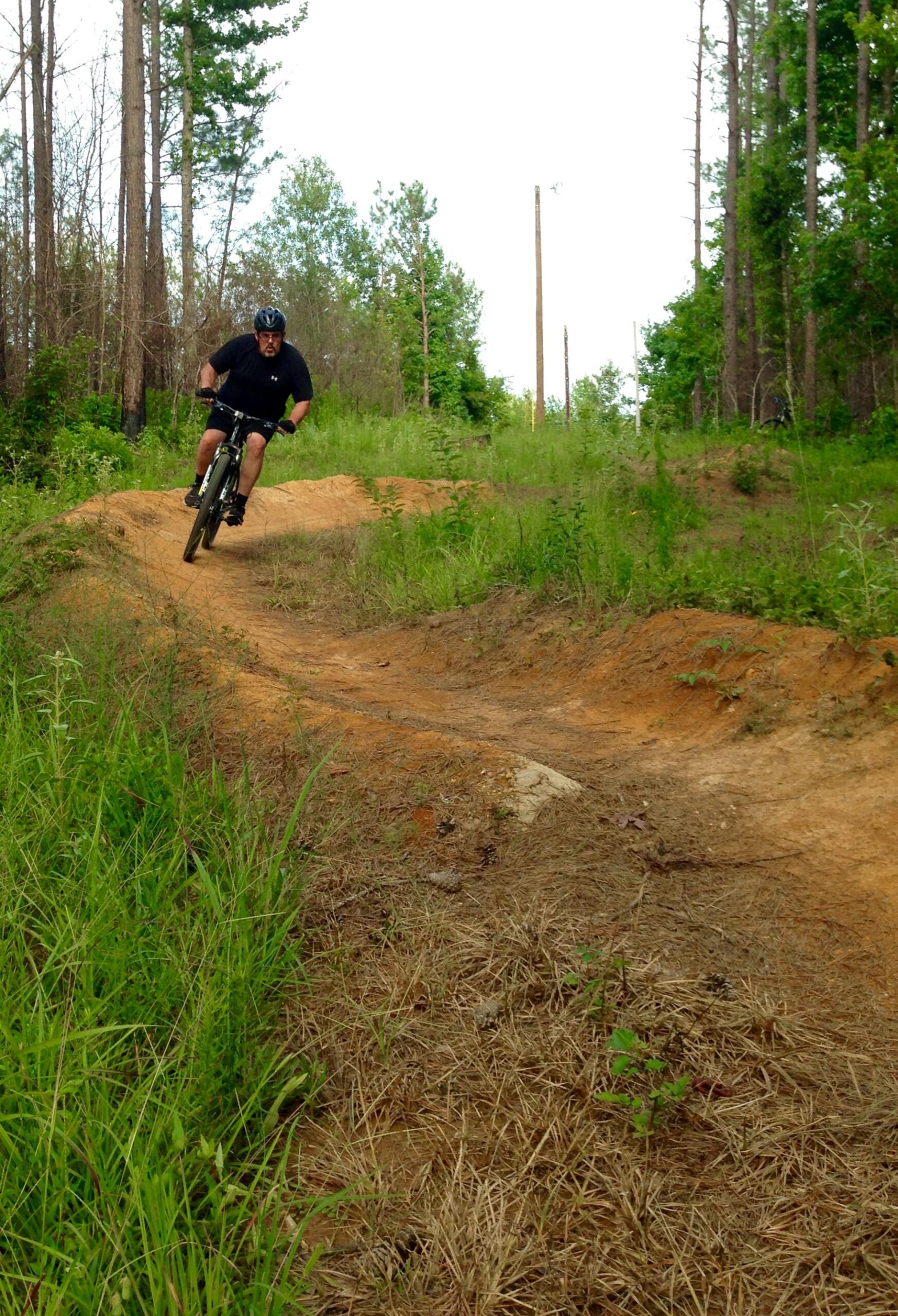 A person riding a mountain bike on a dirt trail surrounded by greenery and trees. The rider is leaning into a turn, showcasing an action pose, with a path of brown earth and grass visible on either side. The background features tall trees and a clear sky. Mt. Zion Bike Trails mountain bike trail.