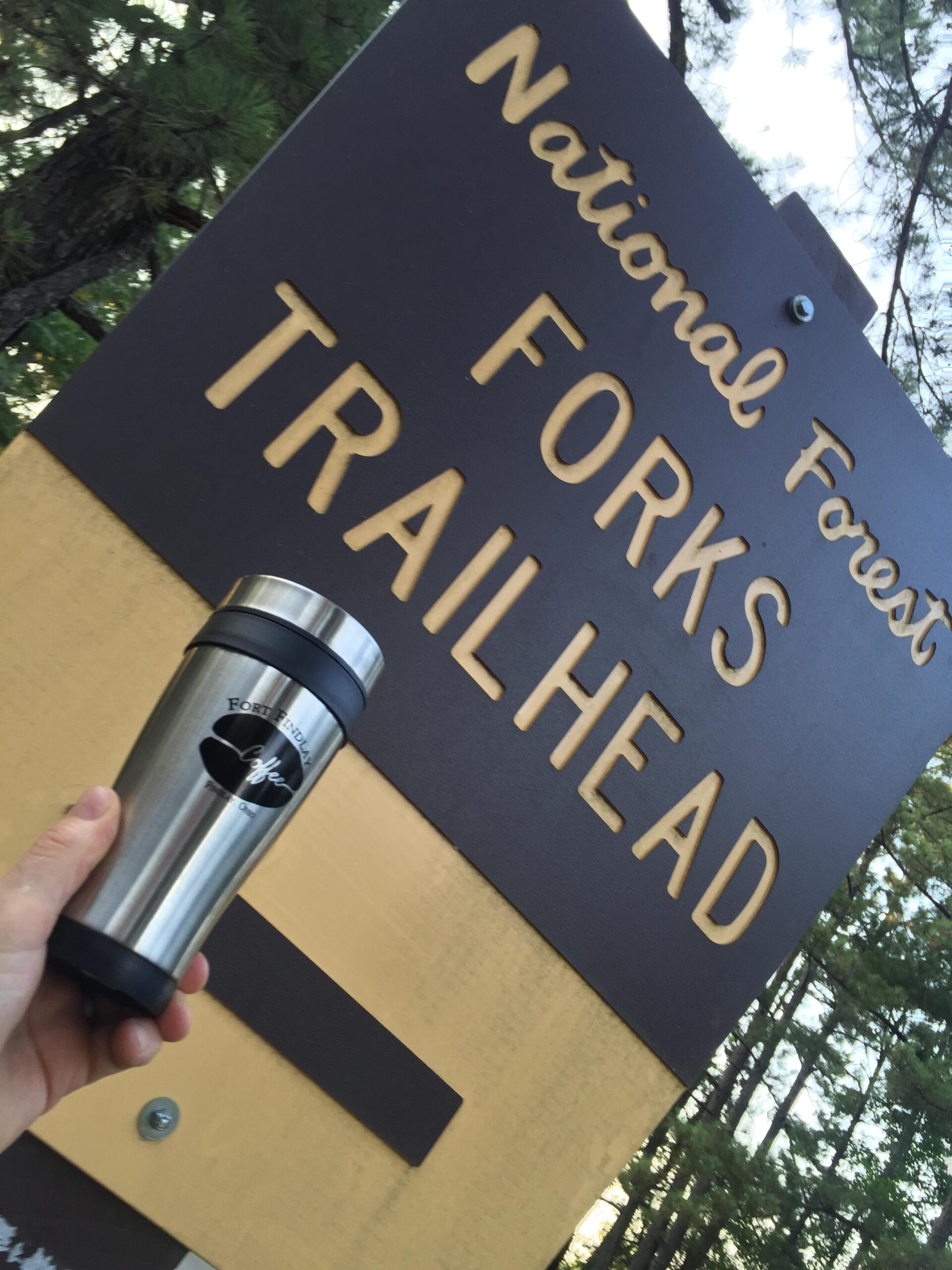 A hand holding a stainless steel travel mug in front of a sign that reads "National Forest Forks Trailhead," surrounded by trees. Forks Area Trail System (FATS) mountain bike trail.