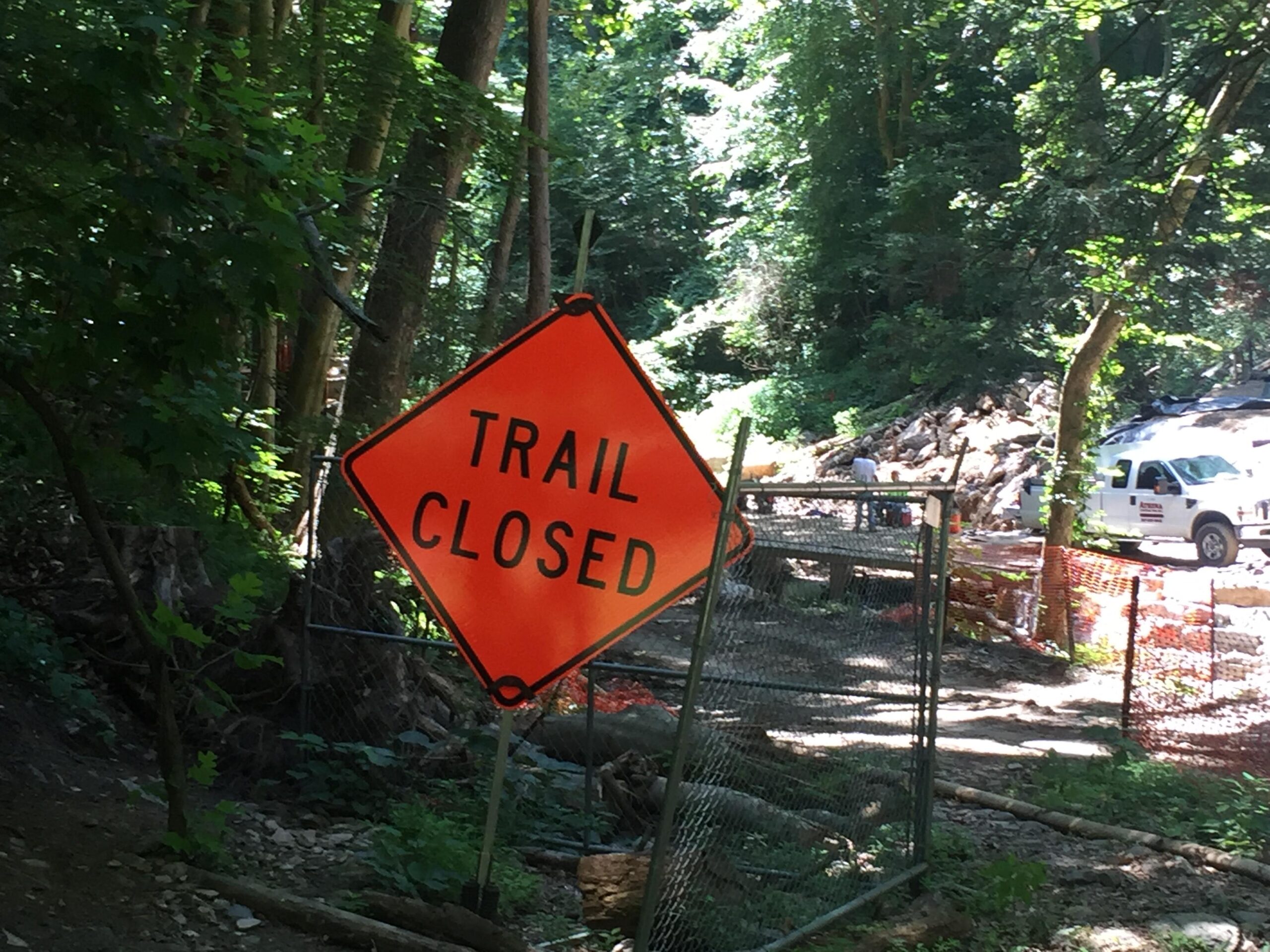 Orange "Trail Closed" sign in a wooded area with construction activity in the background, surrounded by green foliage and fencing. Wissahickon Valley Park mountain bike trail.
