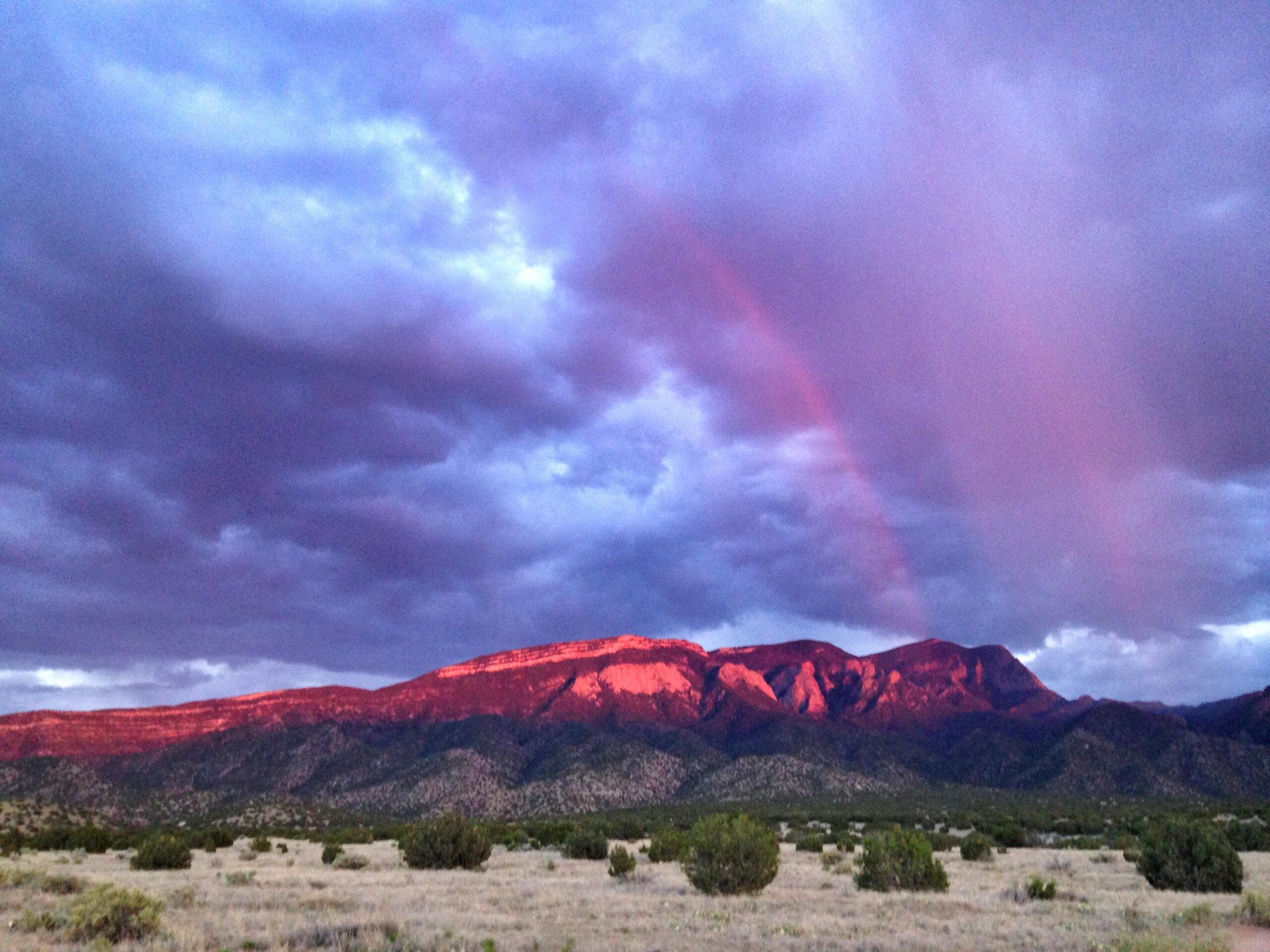 A vibrant landscape featuring dramatic purple and pink clouds above a rugged mountain range, illuminated by the setting sun. A faint rainbow arcs through the sky, adding a touch of color to the serene scene. Sparse vegetation and a gentle slope of land are visible in the foreground. MEAT GRINDER mountain bike trail.