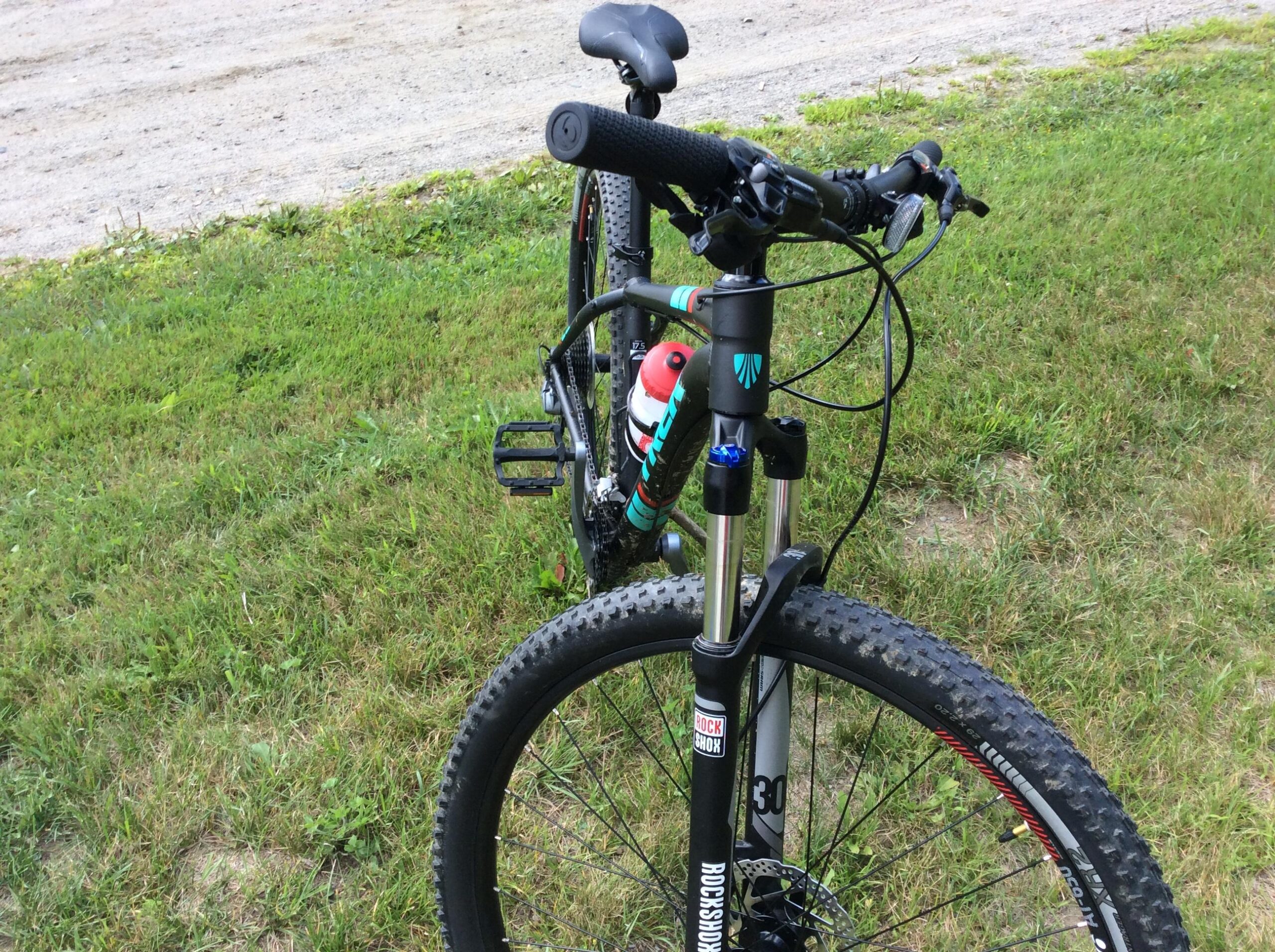 Trek X-Caliber 7: A close-up view of a mountain bike parked on grass. The image focuses on the front of the bike, highlighting the handlebars, tire, and suspension fork. A water bottle is attached to the frame, and the background features a gravel path.