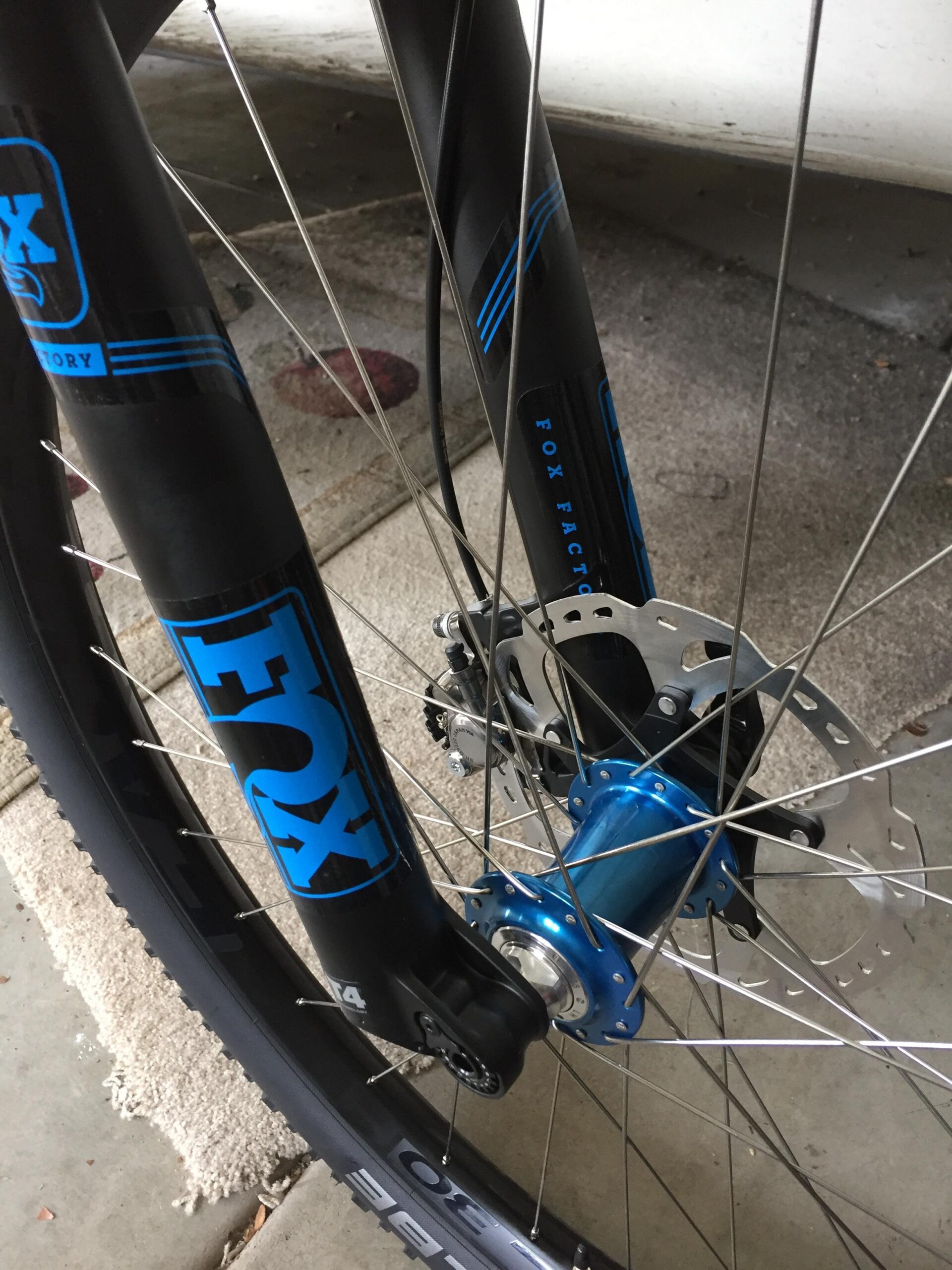 Close-up of a bicycle front fork with black and blue graphics, featuring a disc brake and a blue hub. The image shows the wheel spokes and part of the tire on a concrete surface. Granite Mountain Loop mountain bike trail.