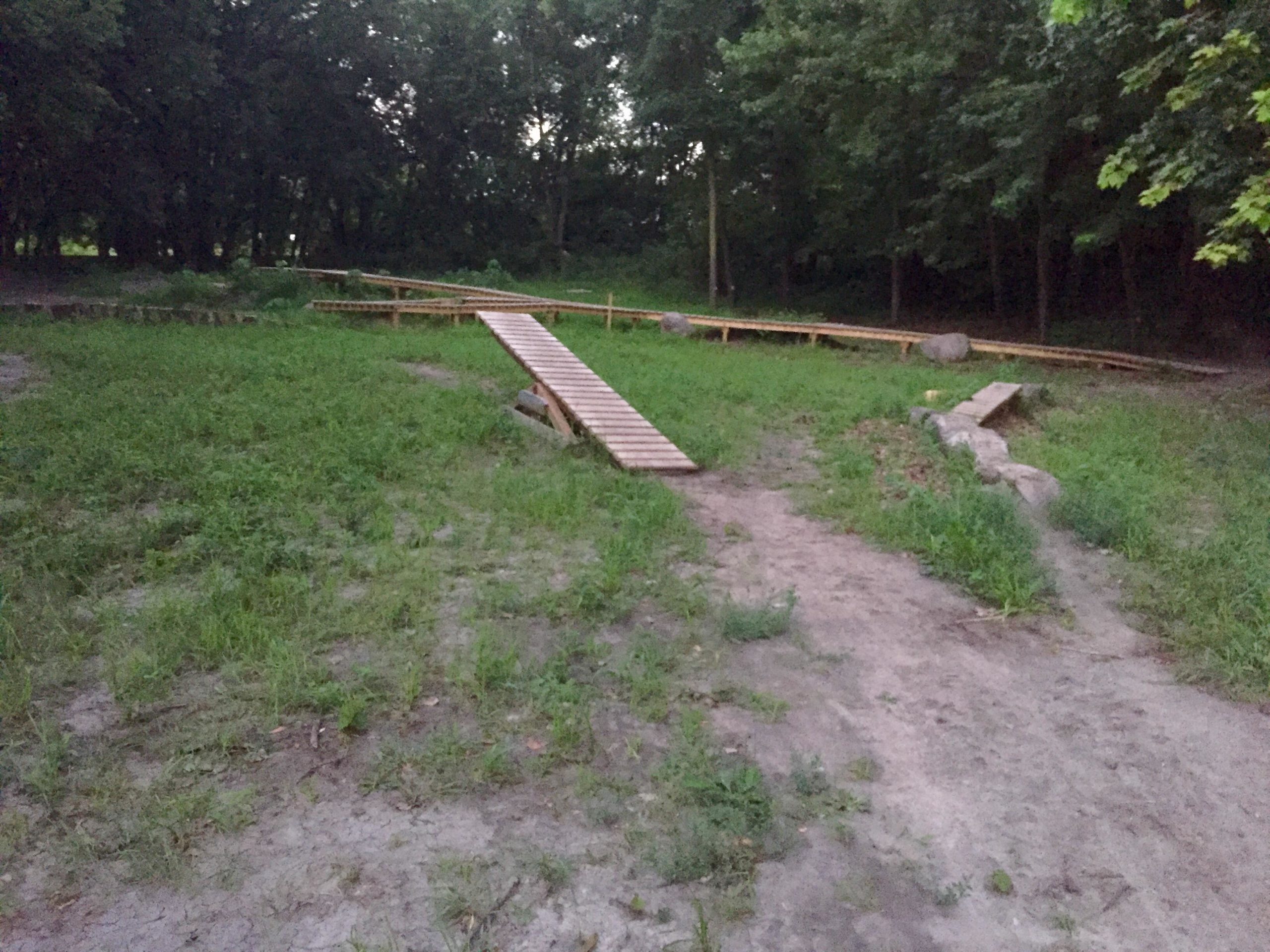 A dirt area in a park with a wooden ramp leading up to a raised platform, surrounded by grass and trees in the background, during dusk. Kiwanis - Mankato mountain bike trail.