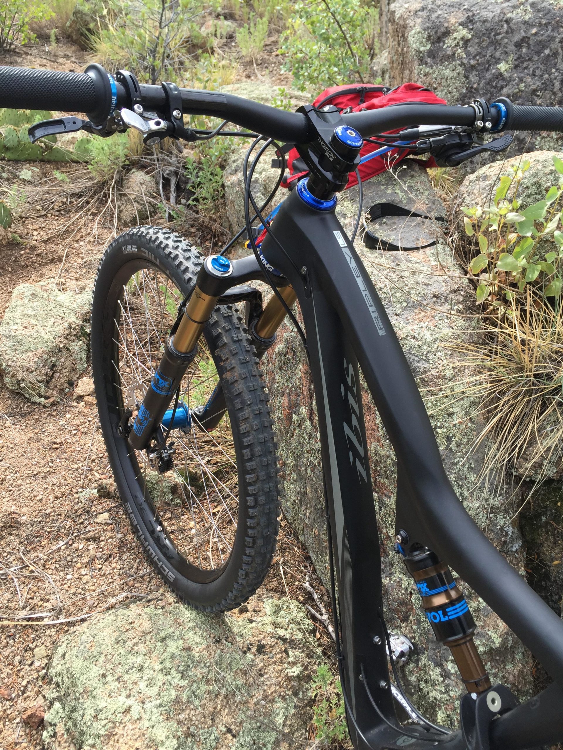 A close-up view of a mountain bike parked on rocky terrain, featuring a sleek black frame with blue and gold accents. The handlebars, equipped with grips and brake levers, have adjustable components visible. The bike's front wheel is angled against a boulder, surrounded by natural vegetation and gravel. A red backpack is partially visible in the background. Granite Basin Loops mountain bike trail.