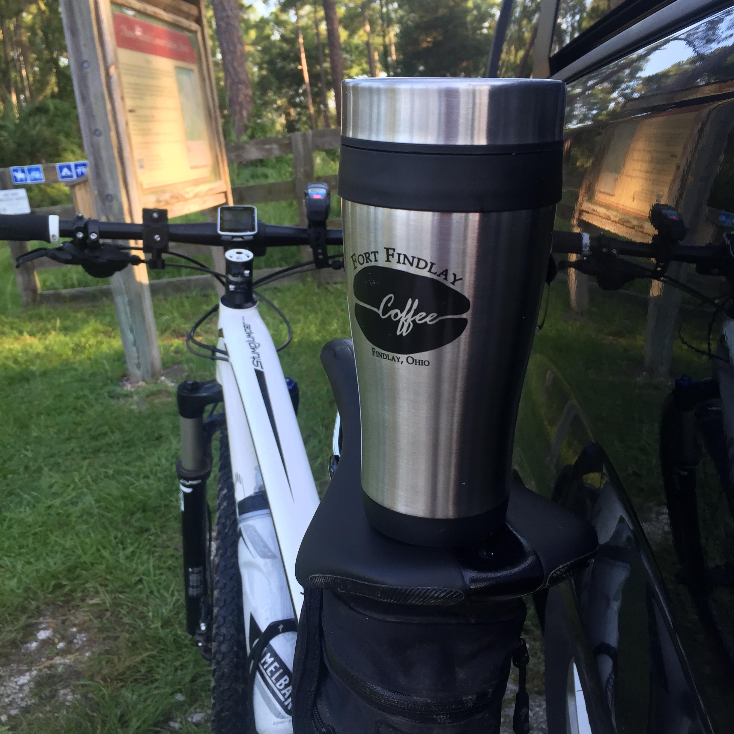 A close-up view of a stainless steel travel mug labeled "Fort Findlay Coffee, Findlay, Ohio," resting on a bicycle rack. In the background, a bicycle with a white frame and visible accessories is parked near a grassy area, with a wooden sign post partially visible. Moses Creek mountain bike trail.