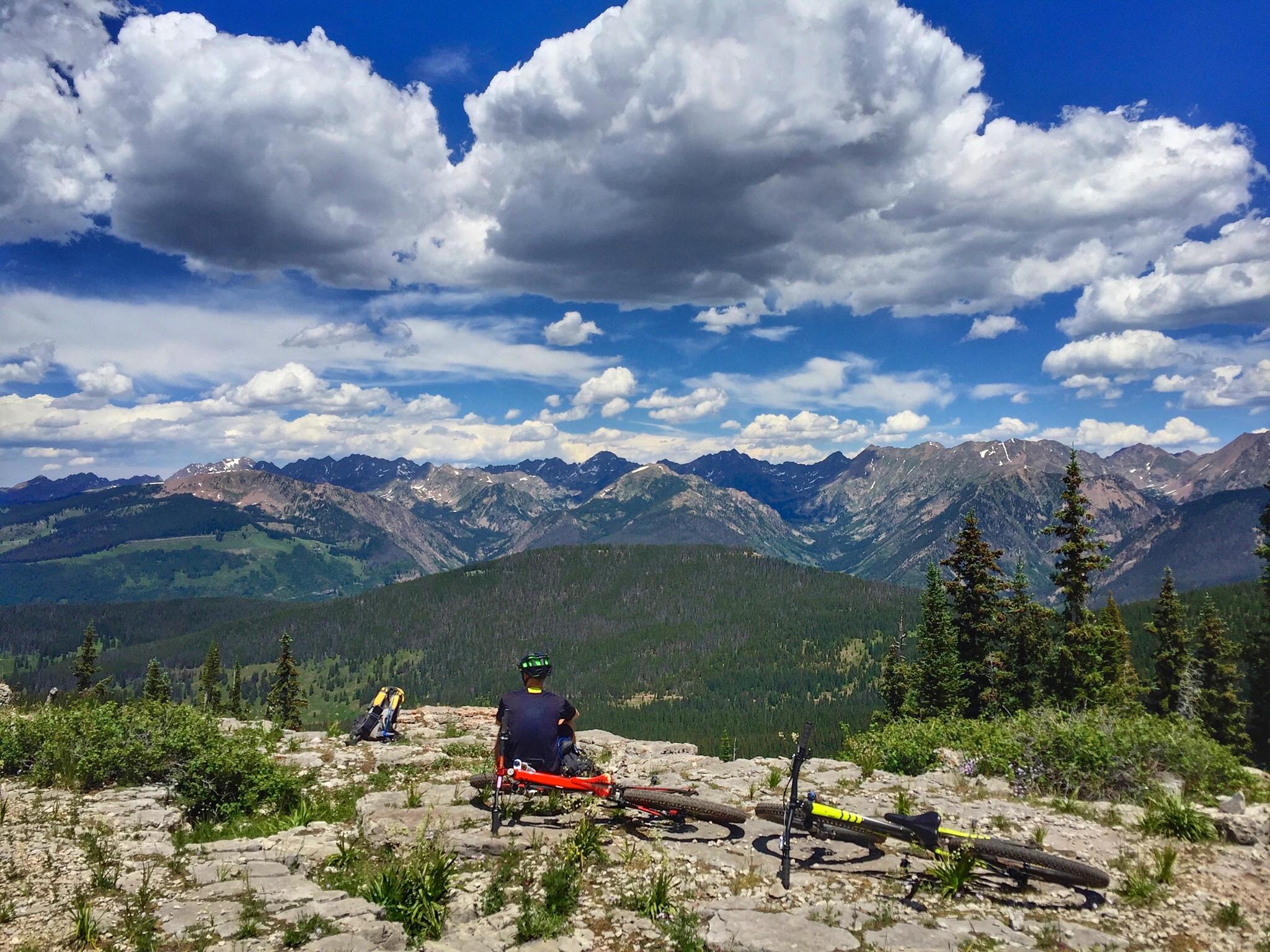 A mountain biker sitting on a rocky ledge, overlooking a vast mountain landscape with lush greenery and a sky filled with white clouds. Two bicycles are resting nearby, with various mountain peaks visible in the distance. Vail Mountain Bike Park mountain bike trail.