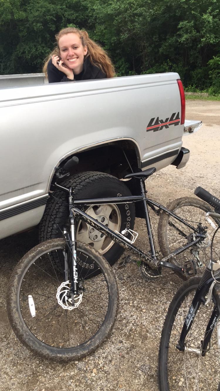 Nashbar AT-2: A smiling young woman sitting in the back of a silver pickup truck, holding a phone, with a mountain bike leaning against the truck's tire on a gravel road. Lush greenery is visible in the background.