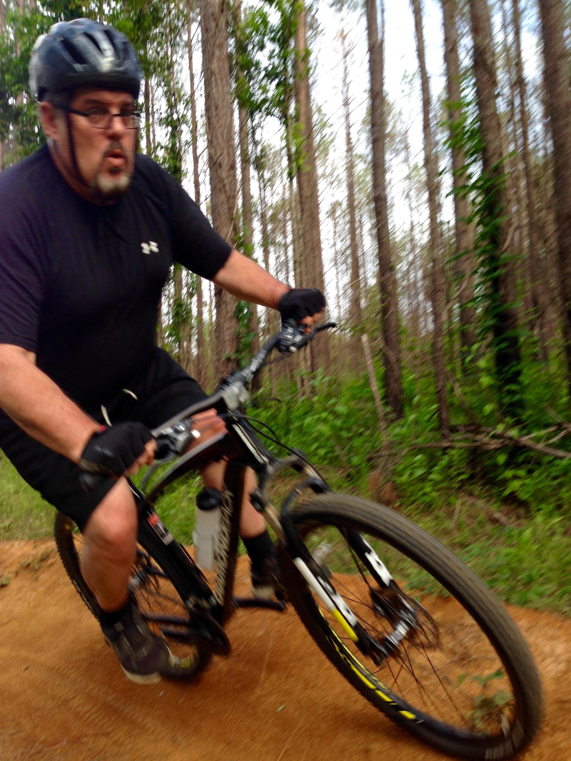 A person wearing a black helmet and gloves rides a mountain bike on a dirt path surrounded by tall trees. The rider is in a dynamic pose, suggesting motion as they navigate the trail. Green foliage is visible in the background, indicating a natural, outdoor setting. Mt. Zion Bike Trails mountain bike trail.