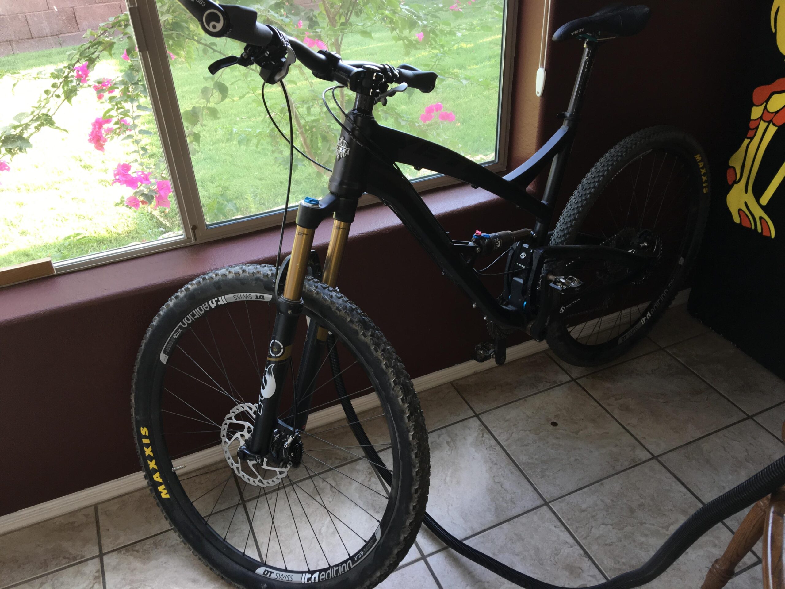 Yeti SB95: A black mountain bike is leaning against a wall near a window, with colorful flowers visible outside. The bike features a front suspension fork, thick tires, and a disc brake. A vacuum hose is resting on the floor beside it. The indoor setting includes tiled flooring and a decorative wall rug hanging nearby.