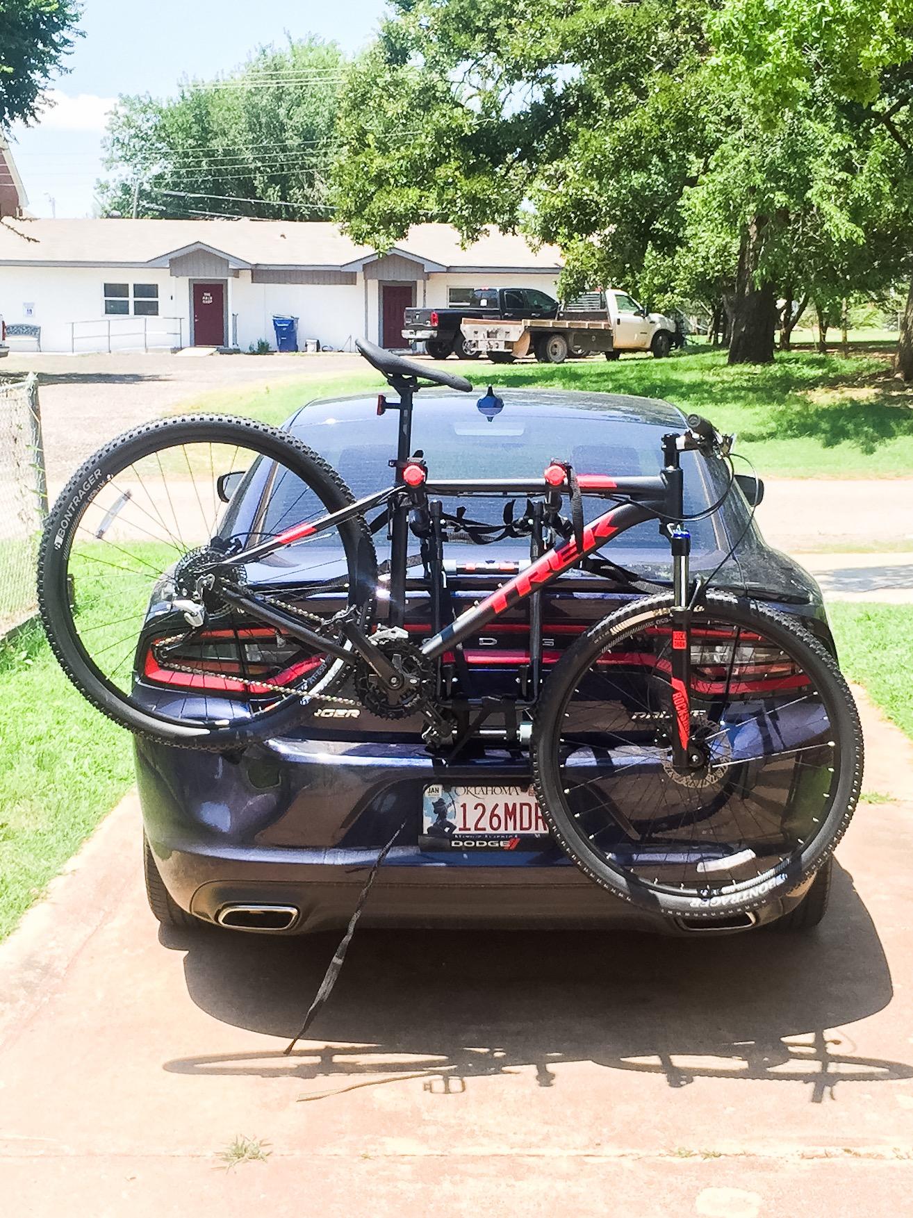Trek Marlin 7: A red and black mountain bike securely mounted on the back of a dark blue Dodge car, parked on a driveway. In the background, a residential area with several houses is visible, along with a pickup truck. Sunlight casts shadows around the scene, indicating a bright day.