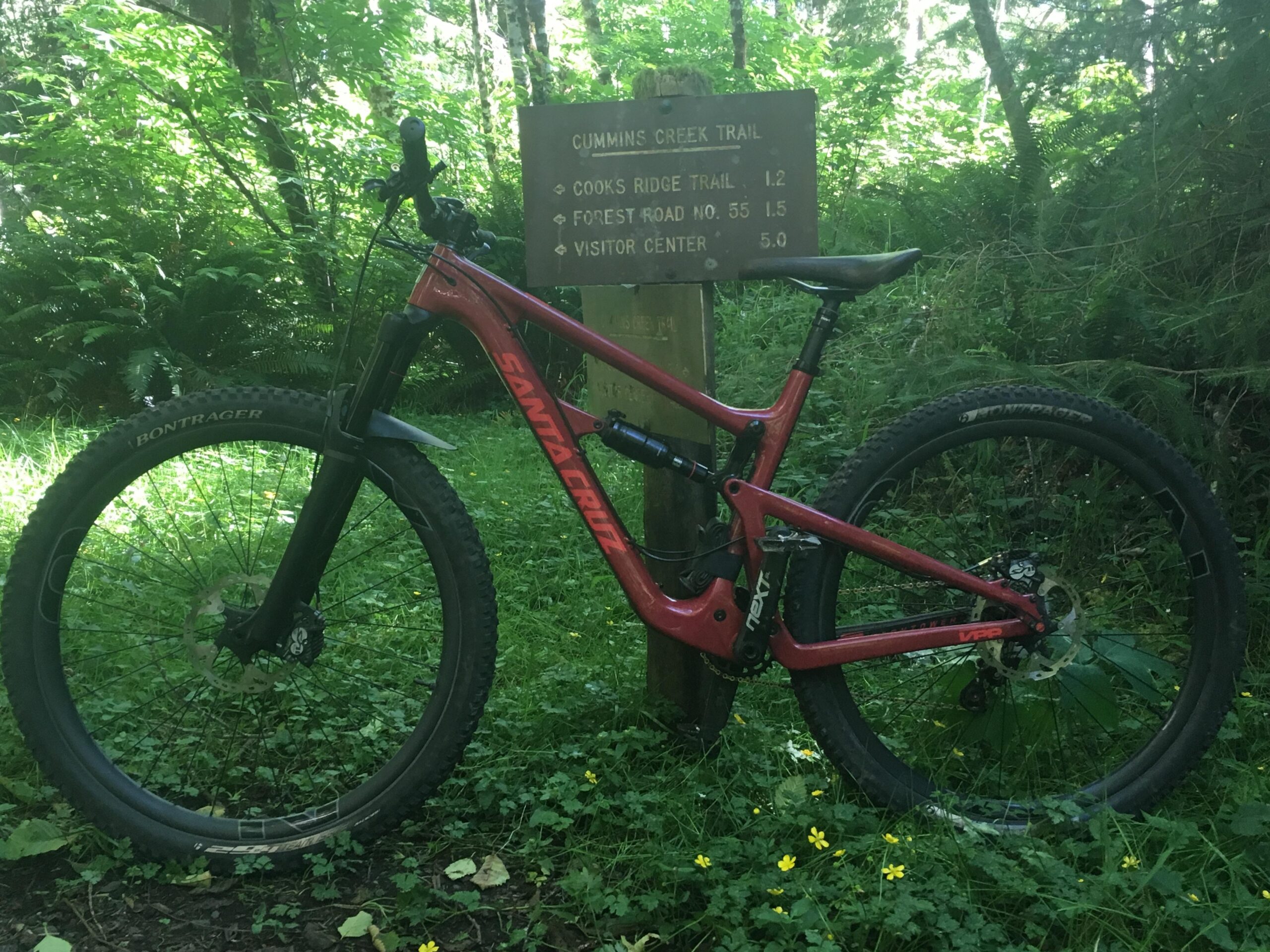 Santa Cruz Hightower 29er/27.5: A red mountain bike resting beside a wooden trail sign in a lush, green forest. The sign lists directions and distances to various trails, including Cummins Creek Trail and Cook's Ridge Trail, along with a Visitor Center. Sunlight filters through the trees, highlighting patches of grass and yellow wildflowers in the foreground.