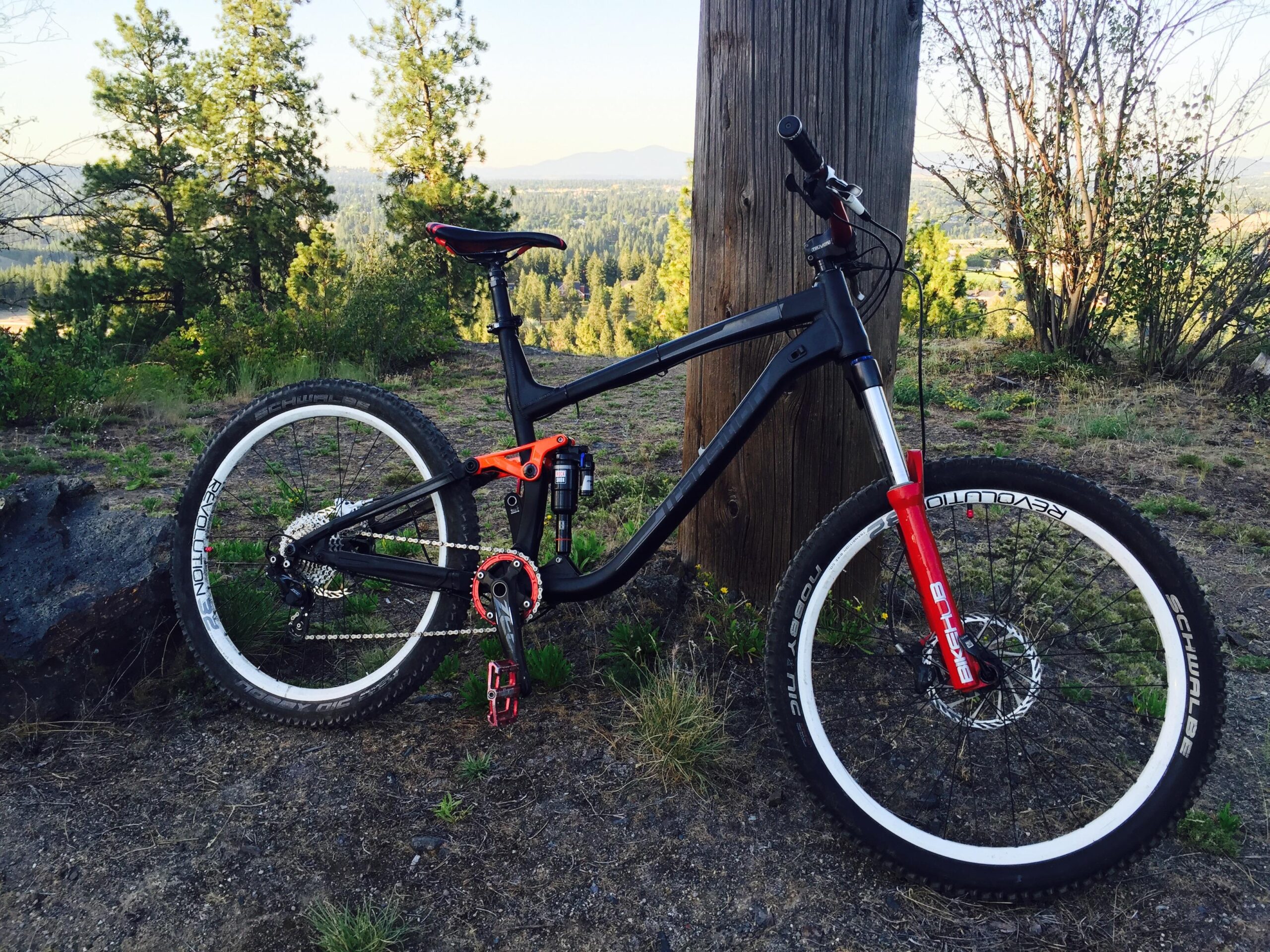 Transition Suppressor: Mountain bike leaning against a wooden post, set in a scenic outdoor environment with green trees and distant mountains in the background. The bike features a black frame with orange accents and red front suspension forks. The ground is sandy with some grass and small plants surrounding the bike.