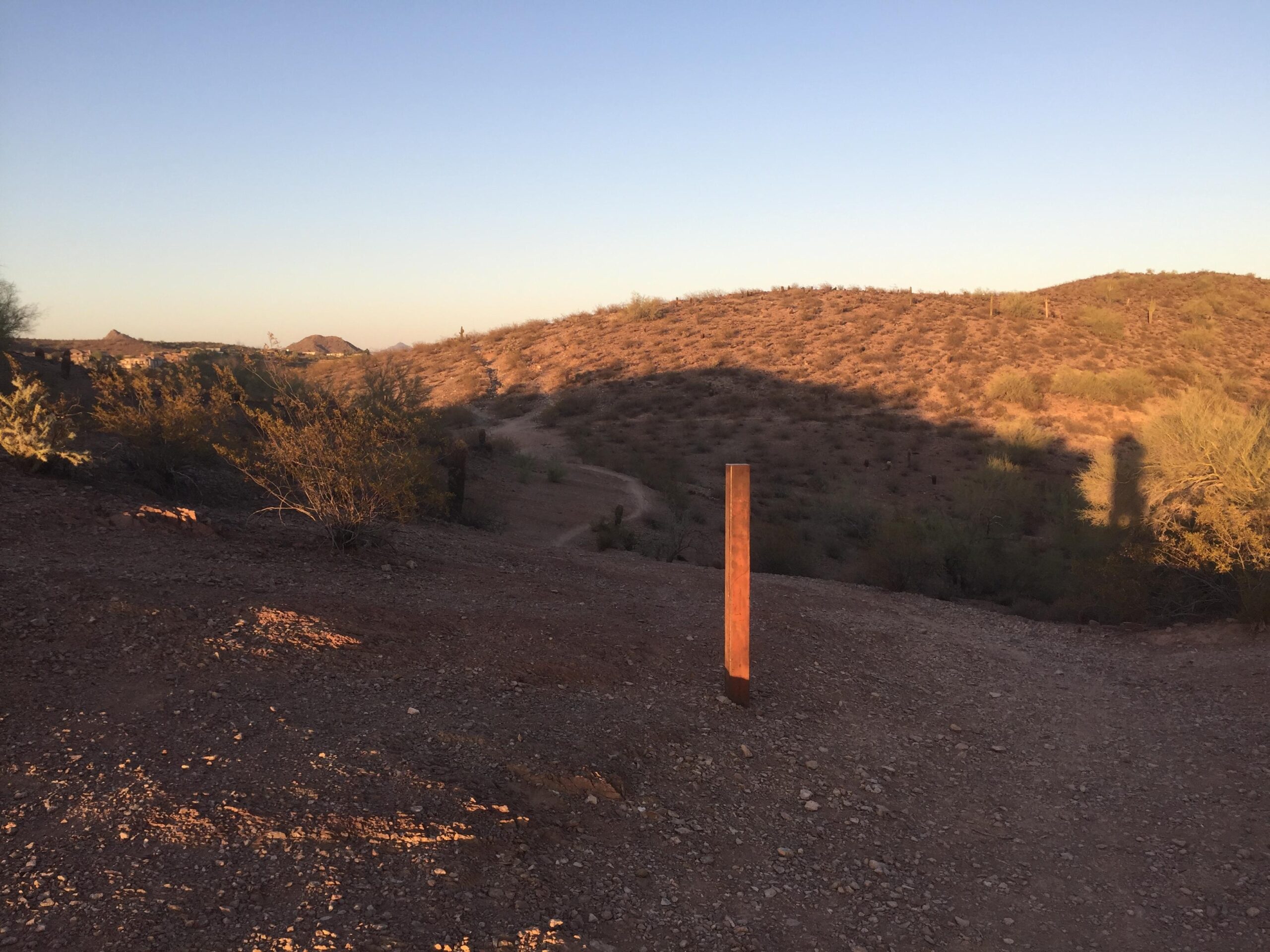 A rugged dirt path winds through a desert landscape at sunset, with rolling hills in the background. A vertical wooden post stands along the path, surrounded by sparse vegetation and rocky terrain, with soft shadows stretching across the ground. Trail #100 mountain bike trail.