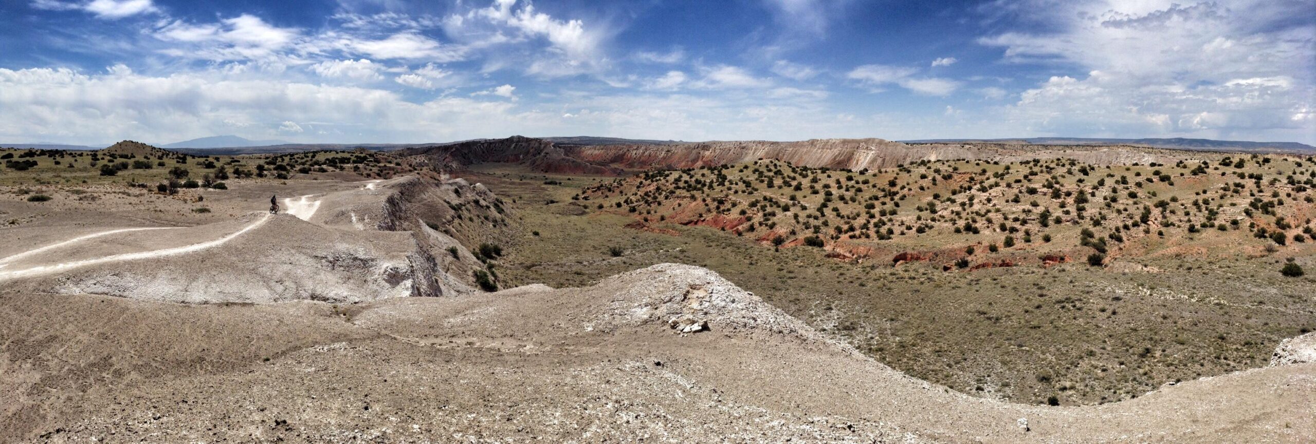 A panoramic view of a rugged, arid landscape featuring rolling hills and sparse vegetation, with a mix of dusty soil and patches of greenery. The sky above is bright with fluffy clouds scattered across a blue background. A cyclist is seen riding along a dirt path that meanders through the terrain, adding a sense of scale to the expansive scene. White Ridge Bike Trails mountain bike trail.