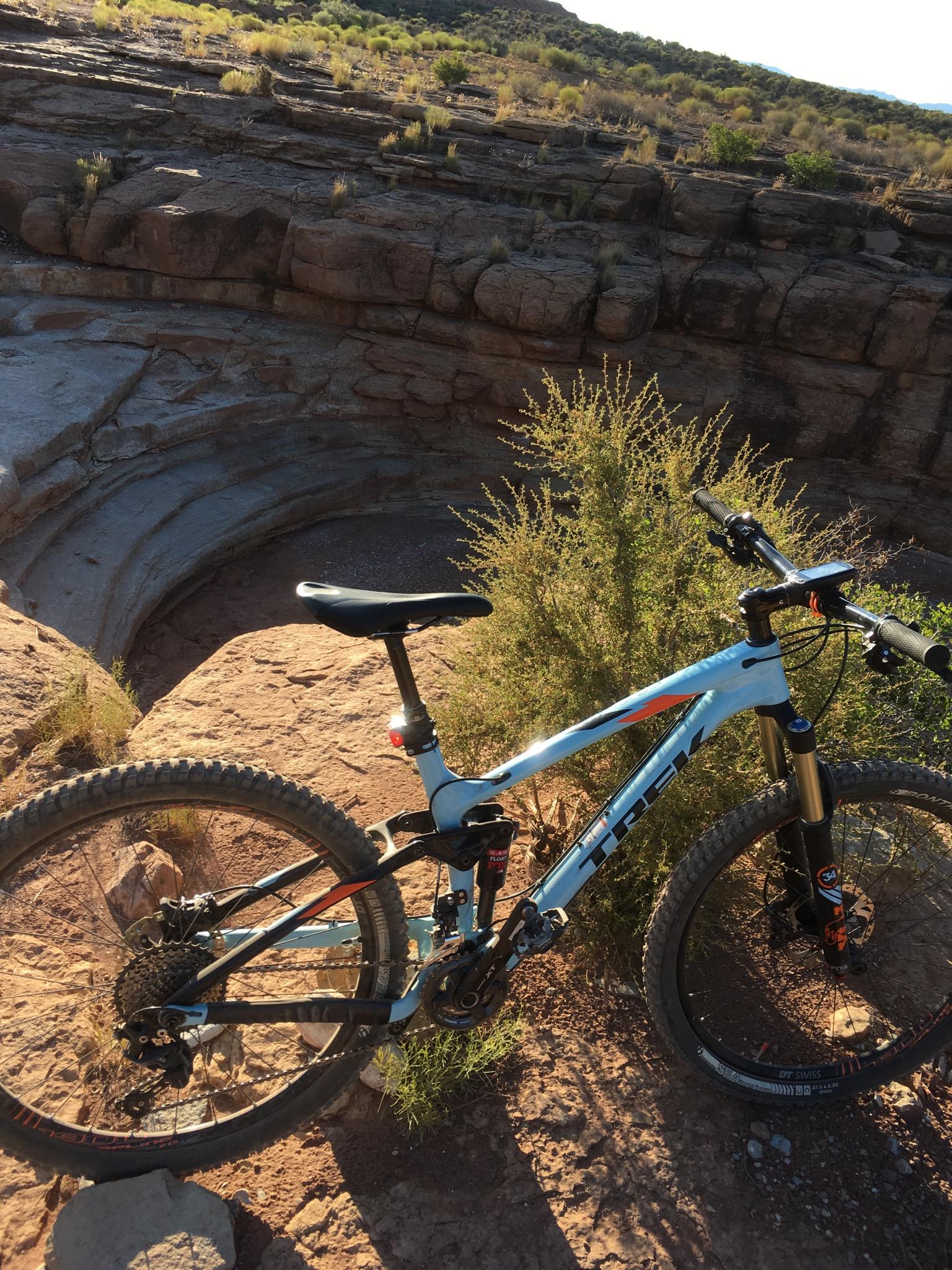 Trek Trek Fuel EX 9.8 27.5: A mountain bike parked on rocky terrain with a circular depression in the background, surrounded by sparse vegetation and a hilly landscape under a clear sky.