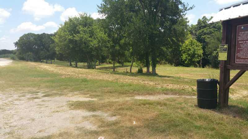 A grassy area with a dirt path leading to a line of trees. To the right, there is a wooden signboard with information and a yellow notice, along with a black trash can nearby. The sky is partly cloudy. Dry Comal Trail mountain bike trail.