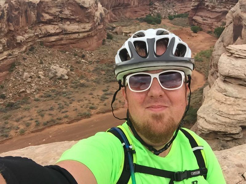 A person wearing a green cycling jersey and a helmet poses for a selfie against a backdrop of rocky terrain and hills. The landscape features reddish-brown rocks and sparse vegetation under a cloudy sky. Moab Brand Trails mountain bike trail.