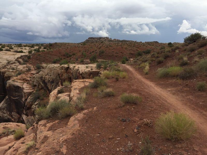 A dirt path winds through a rugged landscape with reddish-brown terrain, featuring shrubs and grasses. In the distance, a rocky outcrop rises against a backdrop of cloudy skies, suggesting an expansive, natural setting. Moab Brand Trails mountain bike trail.