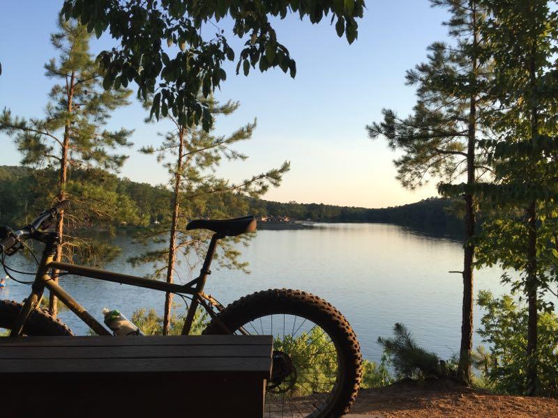 A scenic view of a calm lake surrounded by trees, with a mountain bike resting on a wooden bench in the foreground. The sun is setting, casting a warm glow over the landscape. Oak Mountain State Park Bump Trail mountain bike trail.