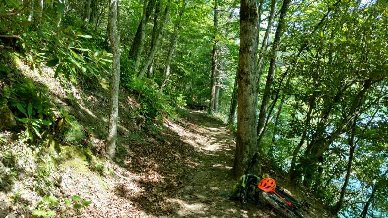 A narrow, winding dirt path through a lush green forest, lined with trees and foliage. A bright orange bicycle helmet rests on the ground beside a mountain bike, partially visible in the foreground. Sunlight filters through the leaves, illuminating the scene with a vibrant, natural feel. Tsali Recreation Area mountain bike trail.