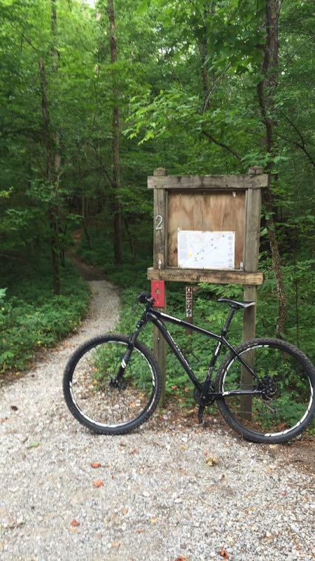 A black mountain bike is leaning against a wooden trail sign marked with the number "2." The sign features a map and trail information. A narrow gravel path winds into a lush green forest filled with trees and dense foliage. The overall scene conveys a sense of adventure and outdoor exploration. Oak Mountain State Park mountain bike trail.