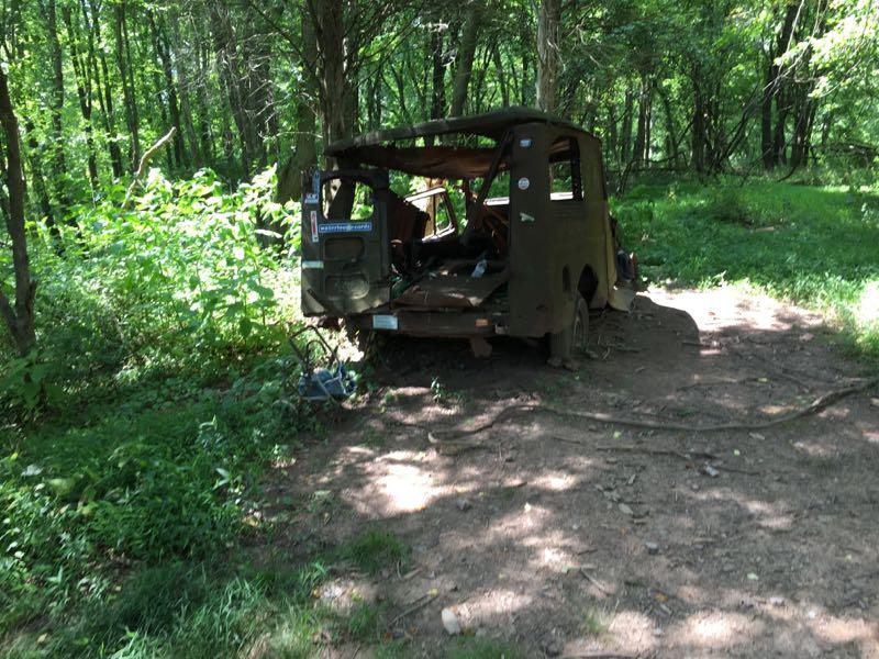 Alt text: An abandoned, rusted vehicle partially hidden in a lush green forest. The vehicle has an open back, showing its dilapidated interior, and is surrounded by dense vegetation and trees, with a dirt path nearby. Schaeffer Farms mountain bike trail.