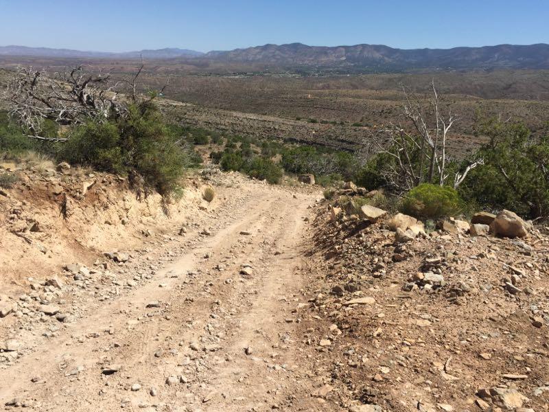 A dirt path winding down a rocky hillside, surrounded by sparse vegetation and distant mountain ranges under a clear blue sky. Dry Canyon (t5574) mountain bike trail.