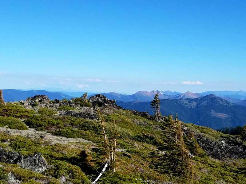 A scenic view of rugged mountains and lush green hills under a clear blue sky. The landscape features rocky outcrops and sparse trees, with distant mountain ranges visible on the horizon. Grayback Trail mountain bike trail.