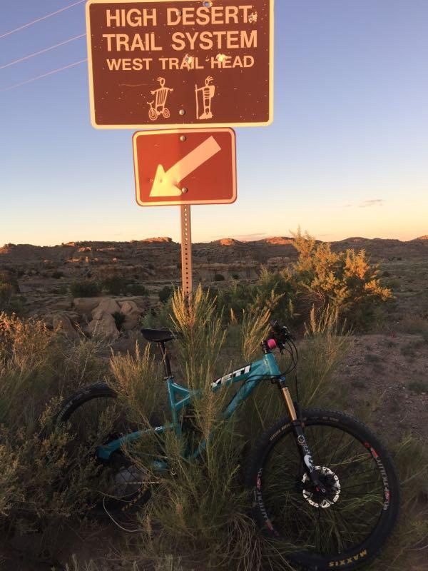 A mountain bike rests beside a sign marking the West Trail Head of the High Desert Trail System, surrounded by desert vegetation. The sun sets in the background, illuminating the landscape with warm tones. High Desert Trail System mountain bike trail.