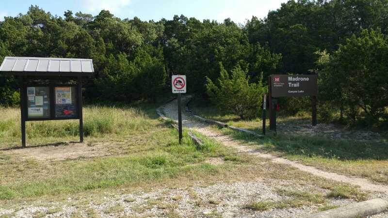 A trail entrance marked by a sign for "Madrone Trail" with surrounding grassy area, a wooden information kiosk, and a "no pets" sign. The path leads into a wooded area filled with trees. Madrone Trail mountain bike trail.