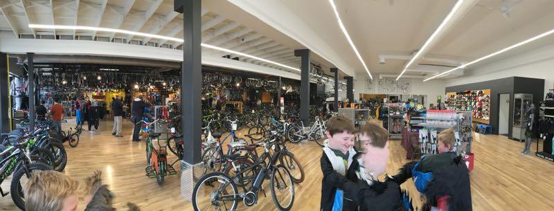 A panoramic view of a large bicycle store featuring numerous bikes displayed throughout the spacious interior. The store has a modern design with bright lighting, wooden flooring, and multiple aisles filled with various cycling gear and accessories. In the foreground, a group of children is seen interacting, while customers browse the store in the background.