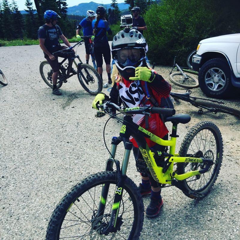 A young child wearing a helmet and protective gear poses with a brightly colored mountain bike on a gravel path. In the background, several adults are gathered, also on mountain bikes, surrounded by trees and mountains in the distance. The child is giving a thumbs-up gesture, indicating excitement. Downieville Downhill mountain bike trail.