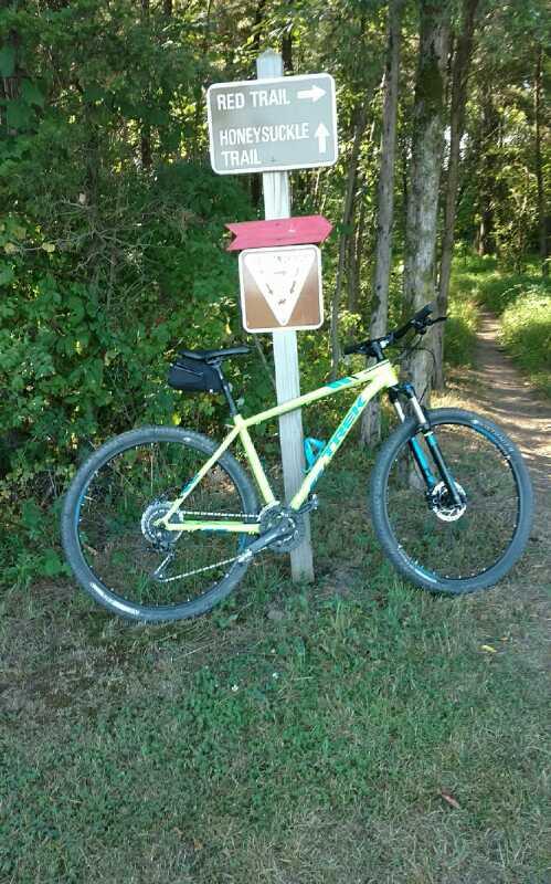 A mountain bike parked beside a trail sign indicating directions for the "Red Trail" and "Honeysuckle Trail" in a wooded area. Green Lane mountain bike trail.