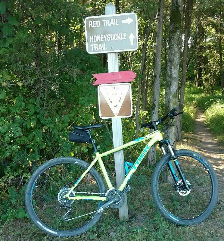 A mountain bike is parked next to a trail sign indicating directions for the Red Trail and Honeysuckle Trail, surrounded by lush greenery and trees. Green Lane mountain bike trail.