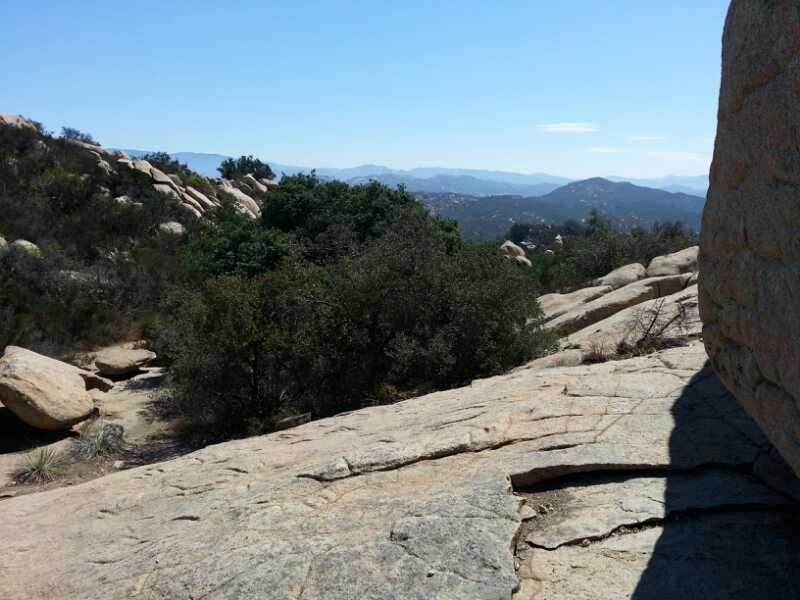 A rocky landscape featuring large boulders and scattered vegetation, with a view of distant mountains under a clear blue sky. The foreground shows a textured rock surface, while the background includes lush trees and rolling hills. Daley Ranch mountain bike trail.