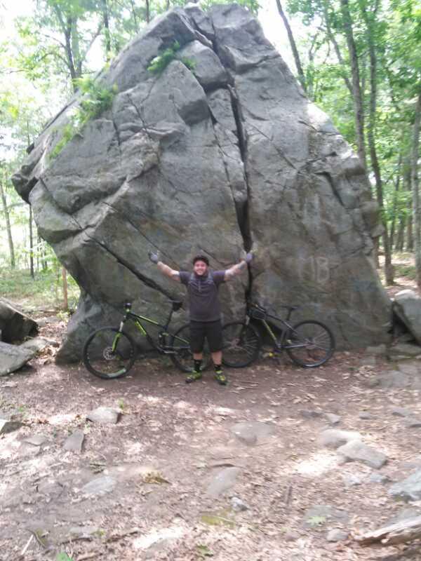 A person standing with arms raised in excitement in front of a large rock formation, with two mountain bikes leaning against the rock. The scene is set in a wooded area with trees in the background and a dirt path visible. Harold Parker State Forest mountain bike trail.