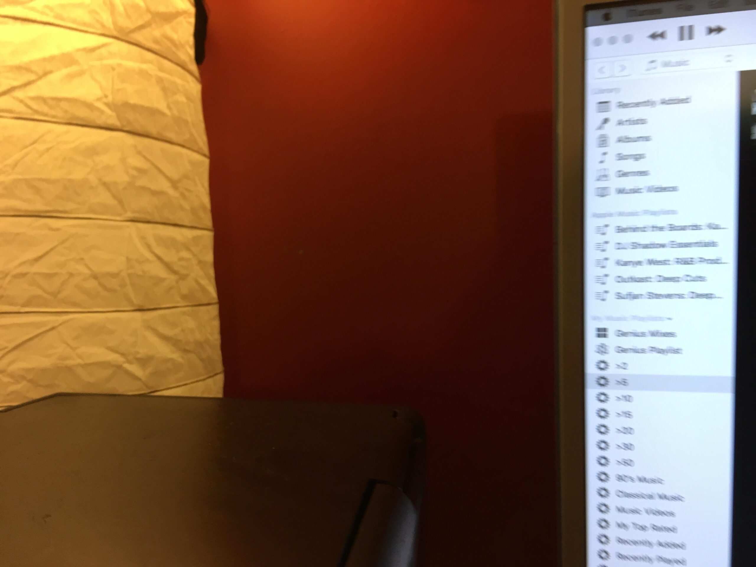 A close-up image showing a desktop corner with a dark surface, a white paper lantern in the background, and a computer screen displaying a music player interface with various playlists and options. The wall behind is painted a warm red color. Yellow River mountain bike trail.