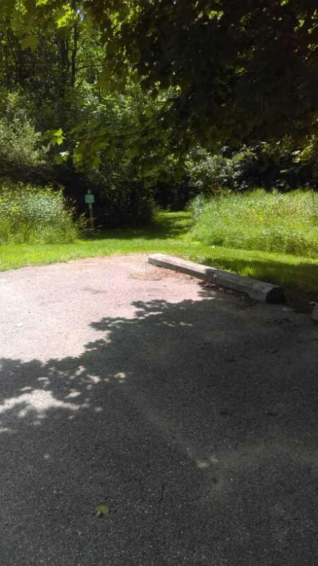 A view of a gravel path leading into a wooded area, bordered by tall green grass and trees. Sunlight filters through the leaves overhead, creating dappled shadows on the ground. In the background, a sign can be seen at the edge of the path, indicating a trail entrance. Seven Lakes State Park mountain bike trail.