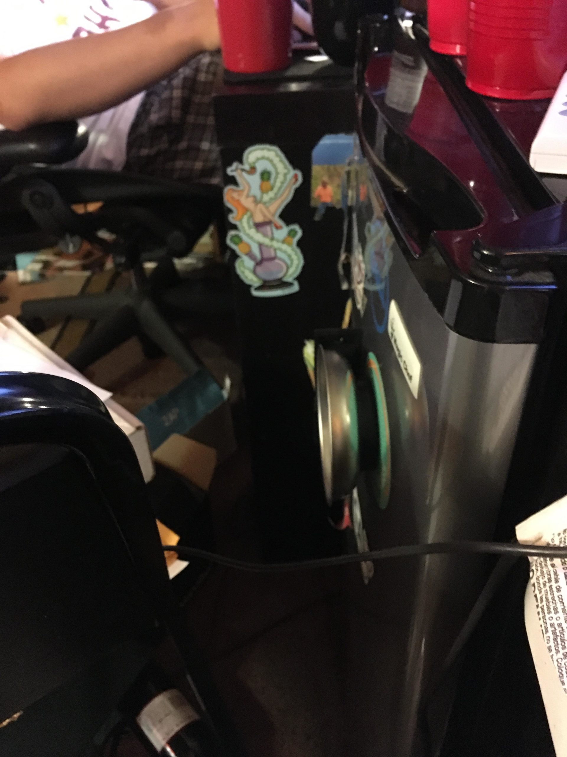 An image showing a cluttered workspace with a person's arm resting on a desk. The desk features a black surface adorned with colorful stickers, including a whimsical design. There are red plastic cups placed on the desk, and a small appliance or container is visible on the right, with a cable running across the surface. The background shows scattered items and additional objects suggesting a lively work or gaming environment. Morningside Nature Preserve mountain bike trail.