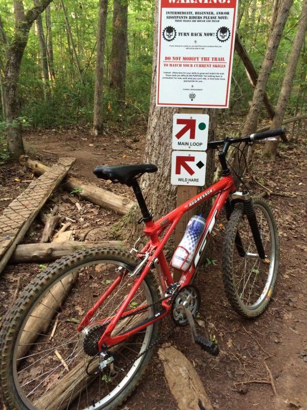 A red mountain bike is leaning against a tree in a wooded area. A warning sign above it advises cyclists to turn back if they are not experienced, and two directional signs indicate the Main Loop and Wild Hare trails. The ground is covered in leaves and dirt, with wooden planks nearby. Fisher Farm Park mountain bike trail.
